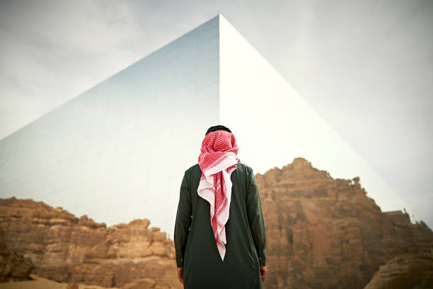 رجل بالشماغ يقف مُدبراً أمام مبنى عاكس مثلّث الشكل وسط تكوينات صخرية في العلا